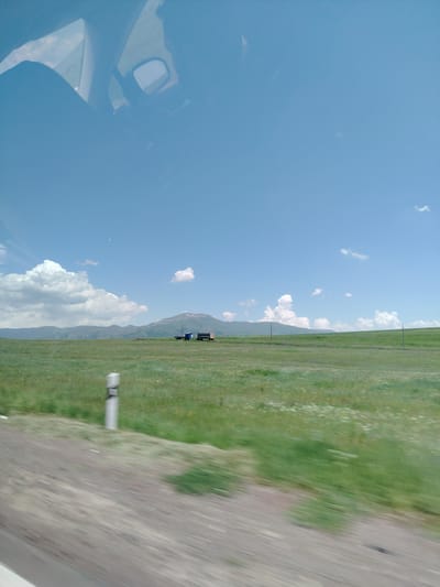 The image shows a landscape scene from inside a vehicle in Hartavan, Armenia. The view is through the windshield and a side window, reflecting the interior of the car, including a rearview mirror and part of the dashboard. Outside, a vast, green, grassy field stretches towards rolling hills and mountains in the distance. The sky is a clear blue with scattered white cumulus clouds. In the mid-ground, on the field, there are two small, dark structures, possibly sheds or small buildings, and a trailer. A white road marker with a black band is visible in the foreground on the left side of the image, indicating the edge of a road. The foreground also shows a blurred area of dirt and gravel, suggesting movement from the vehicle. The overall impression is of a rural, open landscape under a bright, sunny sky.