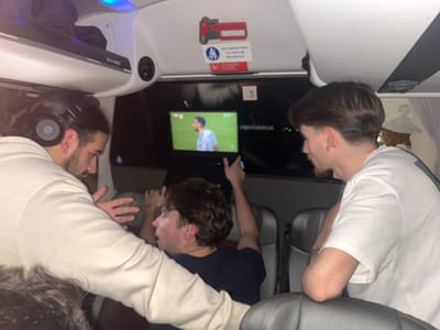 Three men watch a soccer game on a laptop in a bus. The man on the left wears headphones and a light-colored hoodie, leaning over a seated man in a dark shirt. The seated man gestures towards the screen. The man on the right, wearing a white shirt, leans on a seat back, watching the game intently. An "EMERGENCIA" sign is visible above the laptop screen.