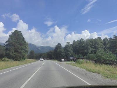 The image shows a two-lane asphalt road with white lane markings, curving gently to the left. The road is bordered by a grassy shoulder on the left and a rocky shoulder with dry grass and vegetation on the right. A white SUV and a dark-colored sedan are traveling in the same direction as the camera on the road. On the left side of the road, a tall pine tree stands prominently, with a dense forest of trees and mountains in the background. On the right side of the road, there is a wooden fence and a sign with Cyrillic text that appears to be "ШЕЛЕСТ БАЗА ОТДЫХА" which translates to "Rustle Recreation Base". The sky is a bright blue with scattered white cumulus clouds. The scene suggests a rural or natural setting, possibly in a mountainous region of Russia, consistent with the location of Turbaza Katun'.