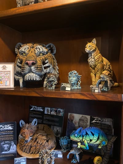 A shelf in a store displays several Edge Sculpture figurines. The top shelf holds a crouching cat and a Medusa head. The bottom shelf features a tiger head, a chameleon, and a curled-up fox. Each item has a tag. There is a brochure on how to create a masterpiece and an ad for the sculpture for the home by Matt Buckley. It can be inferred the location of the image is Llanberis, United Kingdom, based on the text "crafted in England by Matt Buckley".