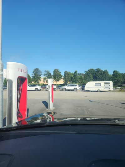 An outdoor Tesla charging station in Strömstad, Sweden, under a clear blue sky features multiple Tesla Supercharger units, the closest one on the left prominently displaying the "TESLA" logo in red. Several electric vehicles are parked at the station, including a white Tesla Model Y and a silver SUV. A white caravan is attached to a silver SUV, parked further back in the lot. Trees and a building line the background. The image is taken from inside a vehicle, with the dashboard and windshield visible in the foreground.
