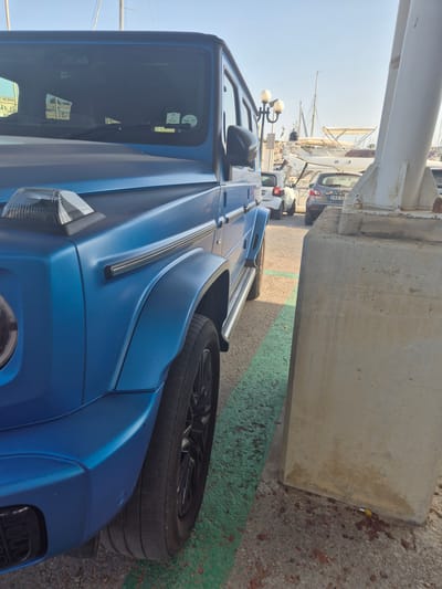 A blue Mercedes-Benz G-Wagen is parked in a marina area, partially obscuring other vehicles and a concrete pillar. The car is matte blue and its front driver-side quarter is prominently featured. A parking sticker is visible on the windshield, indicating "APR 27". In the background, several boats are docked, and the sky is clear and blue. The ground is paved with asphalt and marked with green lines, typical of a parking area. Other cars, including a white city car and a dark gray Nissan Qashqai with license plate "EBS 171", are parked behind the G-Wagen. The scene appears to be located at the Msida Marina, adjacent to Ta' Xbiex in Malta, given the presence of yachts and the characteristic Maltese architecture visible in the distance. The lighting suggests it is daytime, possibly late afternoon. There are no people visible in the image.