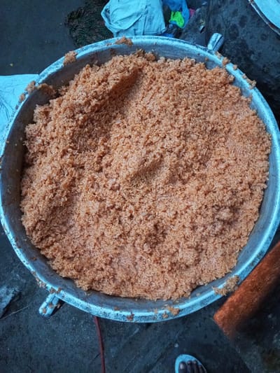 The image shows a large, shallow, aluminum pot filled with orange-colored rice, possibly Jollof rice. The pot has handles. The setting appears to be outdoors, likely Isunba, Nigeria. Fabric, possibly clothing, is seen in the background. A human foot is partially visible in the lower right corner.