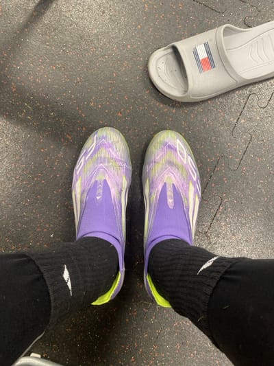 The image displays a downward view of an individual's lower legs and feet on a dark gray, flecked rubberized floor, consistent with an athletic facility or gym in Lawrence Township, United States. The individual wears black athletic pants and black ankle socks, with a white Nike swoosh visible on the left sock. A pair of predominantly purple and lime green athletic shoes with abstract patterns, identified as Adidas, are on the feet. To the upper right, a single light gray slide sandal, bearing a red, white, and blue Tommy Hilfiger flag logo, rests on the floor next to the left shoe. The floor shows minor debris and exhibits interlocking mat edges. No other individuals or actions are depicted.