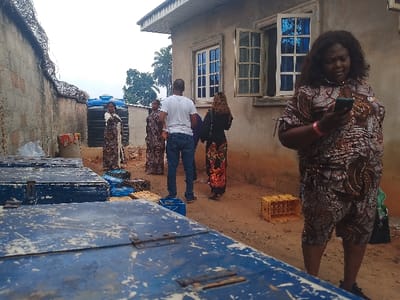 This image depicts an outdoor scene in the city of Ufuma, Nigeria, characterized by a dirt ground and a mix of domestic and storage elements.

In the foreground, several large, rectangular, weathered blue metal boxes with visible hinges and rust are present, one prominently occupying the lower left. To the right, a heavy-set female individual with dark, wavy hair, dressed in a patterned brown and black top and shorts, stands facing left, engrossed in a black smartphone held in both hands. A pink wristband is visible on her left wrist.

In the midground, from left to right, a concrete wall topped with concertina wire runs along the left edge of the frame. Adjacent to the wall, two large blue plastic water storage tanks are visible. Near these tanks, a female individual in a patterned long dress stands, facing right. To her right, a male individual with short hair, wearing a white t-shirt and blue jeans, stands with his back to the viewer, facing towards the water tanks, possibly elevated on a blue plastic crate. Further right and slightly behind him, another female individual with long, curly hair, in a black top and a patterned long skirt (featuring red, orange, and black motifs), faces away from the viewer towards a building. Behind this group, small blue and yellow plastic crates, some containing bottles, are scattered on the ground.

On the right side of the image, a light-colored concrete building with a simple overhang is visible. It features multiple windows with white frames and blue-tinted glass, all open outwards. The sky above is overcast. The ground throughout the scene is unpaved dirt.