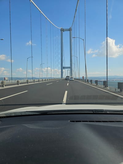 The image shows a view from inside a vehicle driving on a suspension bridge, likely the Osman Gazi Bridge, which spans the Gulf of Izmit and connects Gebze to Orhangazi in the Dilovası region of Turkey. The bridge features multiple lanes of traffic, marked with white lines. Tall, slender lampposts line the sides of the bridge. In the distance, a clear blue sky with scattered white clouds is visible. The water of the gulf is visible on either side of the bridge, with hills or mountains in the far distance. Two white cars are seen driving ahead on the bridge. The foreground of the image is dominated by the interior of the vehicle, including the dashboard and windshield.