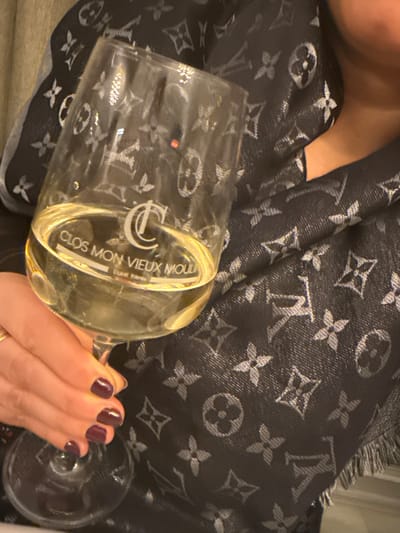 A person holds a partially filled wine glass with "CLOS MON VIEUX MOULIN" inscribed on it. The background features a patterned fabric, possibly a scarf or shawl, with a repeating design of floral-like symbols and what appears to be the letters "LV", commonly associated with Louis Vuitton. The person's hand is visible, adorned with dark, polished nails. The overall composition suggests a relaxed and possibly luxurious setting.