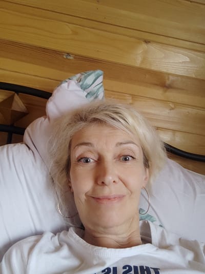 The image shows a selfie of a fair-skinned woman with blonde hair, blue eyes, and fair skin, wearing a white t-shirt. She is lying on a white pillow in what appears to be a bedroom with wooden walls. She is wearing hoop earrings. The background consists of horizontal wooden planks. The overall scene is a close-up of the woman's face and upper body.