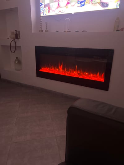 The image shows a living room with a modern electric fireplace set into a white wall. A television is mounted to the left of the fireplace. Decorative candles sit on a shelf below the TV, and a small shelf below the fireplace holds a glass bottle and a dog leash. The floor is tiled. A dark-colored piece of furniture, possibly a couch or chair, is on the right side of the frame.