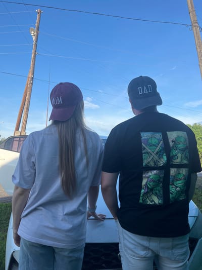 In Mal'tsevo, Russia, a couple stands facing away from the camera, leaning against the hood of a white car under a clear blue sky. The woman on the left wears a maroon baseball cap with "MOM" embroidered in white, and a light-colored t-shirt. The man on the right wears a dark gray baseball cap with "DAD" embroidered in white, and a black t-shirt featuring four panels of the Teenage Mutant Ninja Turtles. They are positioned in an outdoor setting with power poles and overhead wires visible in the background.