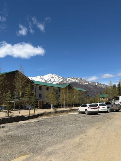 The image displays a scenic outdoor view, likely captured in Denali National Park and Preserve on a clear, sunny day. The sky, a vibrant blue with scattered white cumulus clouds, occupies a significant portion of the frame, indicating pleasant weather.

In the background, majestic snow-capped mountains rise, their rugged peaks partially covered in white snow, suggesting a cool climate or high altitude. The mid-ground features several buildings with prominent dark green, sloped roofs, contrasting with their light-colored walls and numerous windows. These structures appear to be lodges or cabins, possibly offering accommodations within the park. Sparse, thin trees with pale trunks and minimal green foliage are planted around the buildings and along the foreground, hinting at an early spring or late autumn setting.

To the right, a wide gravel road or parking area extends into the foreground. Several vehicles are parked along this gravel surface. A white Dodge Durango SUV is prominently visible, followed by another white van or SUV. Behind these, a silver Chevrolet pickup truck is also parked. On the rear of one of the white vehicles, the visible text "www.denalialaska.com" is a clear location cue.

The scene depicts a tranquil and picturesque environment, characteristic of a national park setting. No people are visible, emphasizing the natural beauty and the quiet atmosphere of the area.