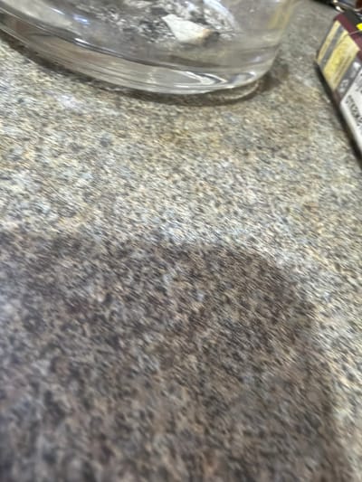 The image shows a close-up view of a countertop with a mottled grey and brown pattern. In the lower left corner, part of a clear glass object, possibly a dish or lid, is visible. A corner of a product box with yellow and white text is at the top left. The environment appears to be indoors. There is no information in the image to determine the location in Fenland District, United Kingdom. There are no people, actions, or interactions visible.