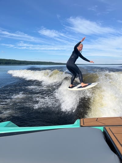 A young woman in a black wetsuit is wake surfing on a body of water, likely a lake, given the treeline in the background. She is positioned on a surfboard with a black and white design, actively balancing and riding a large wave created by a boat, part of which is visible in the foreground. The water is dark and choppy, with white spray from the wave. The sky is blue with wispy white clouds. The location is Gromovo, Russia.