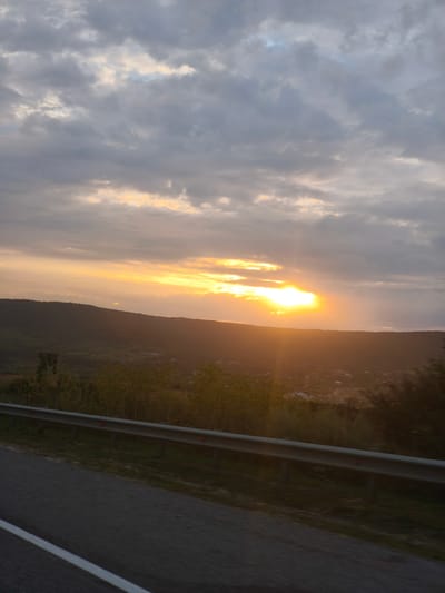 The image depicts a scenic landscape at sunset, likely captured from a moving vehicle on a road. In the foreground, a grey asphalt road with a white line is visible. Adjacent to the road, a metal guardrail with reflective red markers runs horizontally. Beyond the guardrail, a densely vegetated hillside slopes downwards, dotted with small buildings that suggest a rural settlement. The midground is dominated by this hillside and its scattered structures. The background features a vast expanse of sky filled with dramatic clouds. The setting sun casts a warm, golden light, with visible rays of light piercing through the clouds and illuminating the landscape with an orange and yellow hue. The overall impression is one of a tranquil, natural setting bathed in the soft light of dusk. No people or specific actions are discernible in the image. The location is identified as Tatarka, Russia.