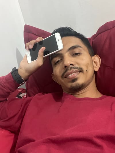 An adult male subject is depicted in a close-up, chest-up view, appearing to be in an indoor setting in Alueduamuka S, Indonesia. The individual has dark hair, a mustache, and a short beard. He is wearing a maroon long-sleeved sweatshirt, and a black wristwatch with a dark strap is present on his left wrist. His left hand is holding a white smartphone with a dark, inactive screen, positioned against his forehead. The subject is looking directly towards the camera with a slight smile. He is leaning against or supported by a dark red cushion, with a plain light-colored wall visible in the upper left background.
