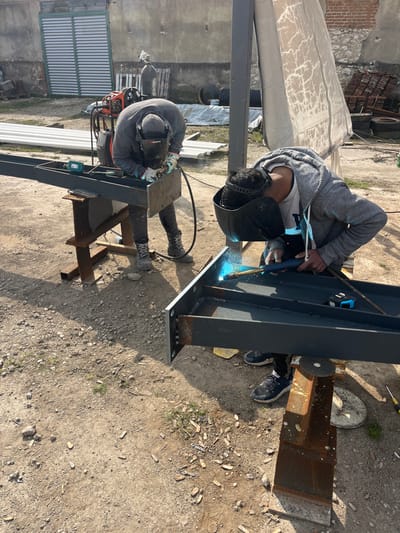 An outdoor scene in Zlatovrah, Bulgaria, depicts two individuals engaged in metal welding activities on an unpaved, dirt and gravel surface.
In the foreground and center-right, a male individual wearing a grey hooded sweatshirt over a white t-shirt, dark trousers, and black sneakers, is positioned kneeling or squatting. He is actively welding a dark grey, I-beam-like metal structure with a visible blue arc and bright light emanating from the welding torch he holds in his gloved hands. His welding helmet is lowered, covering his face. The metal beam he is working on has a bolted end plate and rests on a rusty metal stand. A blue and black measuring tape lies on the beam.
To the left and slightly behind, another male individual, wearing a grey hoodie, dark work trousers, and work boots, is also welding a similar dark grey metal beam. He is bent over, wearing a black Wurth branded welding helmet and work gloves, with sparks visible around the point of welding. The beam he is working on is supported by two rusty metal sawhorses. A teal Makita power tool is visible on this beam.
In the background, an orange welding machine with associated cables is visible. To its left, two gas cylinders (one green, one grey) and wooden pallets are present. Behind the welders, there is a light grey corrugated metal garage door on a building with a textured wall, and various discarded materials including old tires, corrugated metal sheets, and what appears to be a stack of roof tiles are piled. A large, weathered white fabric or tarp hangs from a vertical metal beam on the right side of the background. The ground is dry, with scattered small rocks and sparse patches of weeds. Long shadows are cast from right to left, indicating bright daylight.