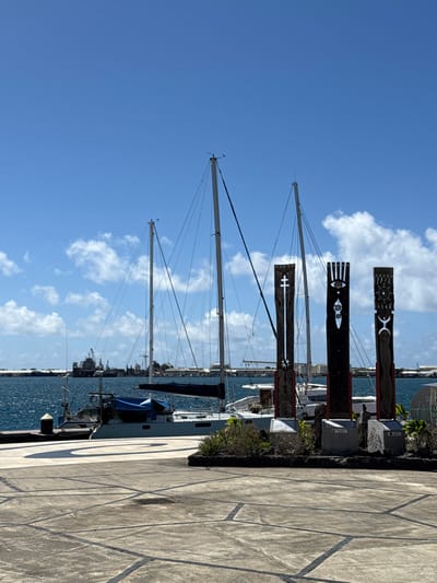 The image depicts a waterfront scene in Papeete, France, under a clear blue sky with scattered white clouds. In the foreground, a paved concrete ground with geometric dark-lined patterns is visible, featuring a large, lighter-colored spiral design to the left. On the right, a raised, dark-colored planter contains low-lying green foliage. Within or behind this planter stand three tall, dark wooden carved pillars, resembling totem poles, each mounted on a grey base. The leftmost pillar displays a white cross-like symbol at its apex and intricate carvings below. The central pillar features a prominent stylized eye at the top, with vertical lines and abstract symbols. The rightmost pillar includes a crescent moon symbol at its peak and detailed vertical carvings.

In the midground, several boats are docked in the blue water of the harbor. A prominent grey catamaran sailboat, equipped with two visible masts and dark rigging, is moored, with its dark blue sail cover deployed. To its left, a smaller vessel, possibly a utility or fishing boat, is also docked, partially covered by blue tarps. A dark bollard is visible on the concrete pier near the boats.

The background across the water reveals an industrial shoreline. Numerous structures, including what appear to be large cranes and industrial vessels, are present at a distant shipyard or port facility. The overall environment is bright and sunny, indicative of daytime. No people are visible in the scene.