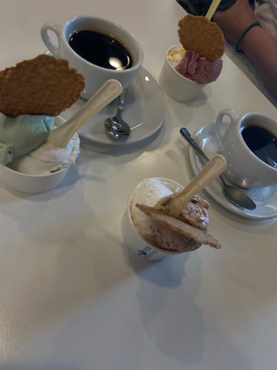 The image captures a table setting featuring three cups of gelato and two cups of coffee, likely taken at a café in Lecco, Italy. Each gelato cup contains two scoops of different flavors, topped with a wafer cookie and a plastic spoon. The coffee cups are white ceramic and sit on matching saucers, each with a spoon. The table is white. In the upper left corner of the image, part of a person's arm is visible. The overall composition suggests a casual cafe setting.