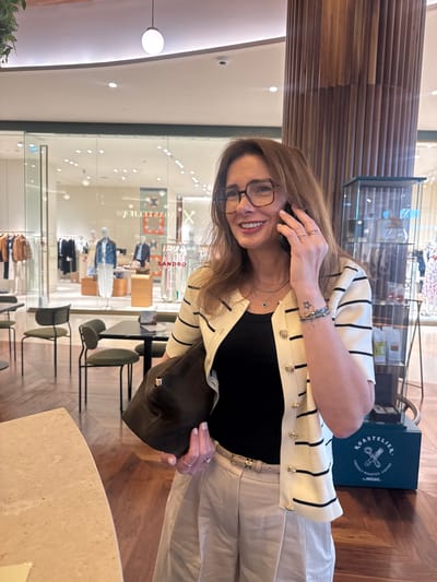The image shows a woman indoors, possibly in a retail environment. She is wearing glasses, a black top, a white cardigan with black stripes, and light-colored pants. She holds a dark-colored hat or bag in one hand and appears to be on a phone call. In the background is a store with clothing displays visible through a glass window. The brand "SANDRO" is visible on the window. Furniture, including chairs, is also visible. A wooden column and a display case with a product box that reads "ROASTELIER COFFEE" can be seen as well. The setting appears to be a modern retail space. The location is Senhora da Hora, Portugal.