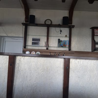 The image shows an interior view of a structure with a textured, light-colored wall and a wooden beam ceiling. A wooden shelf unit with glass shelves is prominent in the center. On the top shelf, two black speakers and a round analog clock are visible. The glass shelves below hold various items including what appear to be small boxes of juice or drinks, a small decorative object, and a stack of what looks like electrical switch plates. Below the glass shelves, another wooden shelf has several circular coasters or lids with colorful designs. The bottom portion of the image reveals a textured, light-colored wall surface with vertical dark wooden posts. The overall impression is that of a bar or service counter area. The scene is located in the city of Dubrivka, Ukraine.