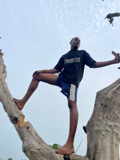 A young man is climbing on a large, fallen tree branch in Agba Umana, Nigeria. He wears a black t-shirt with white text, blue shorts with a white stripe, and is barefoot. The background is a light-colored sky. The image is taken from a low angle, looking up at the man.