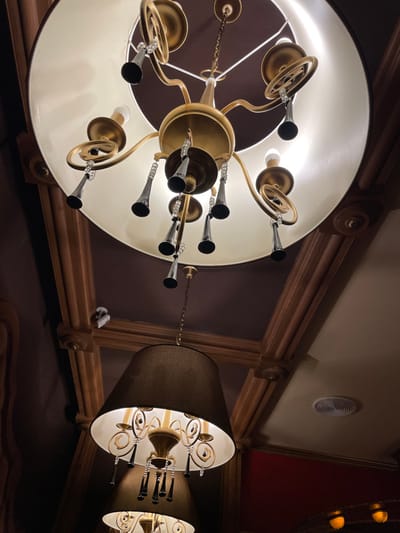The low-angle shot captures an opulent interior featuring two prominent chandeliers. The larger one, on the left, boasts a round, illuminated shade with ornate gold fixtures extending outward, each culminating in a trumpet-like light. A second, smaller chandelier with a cylindrical shade and similar gold accents is visible on the right. Decorative wooden beams and architectural moldings frame the scene, hinting at a classical or historic setting. An air vent is visible on the ceiling.