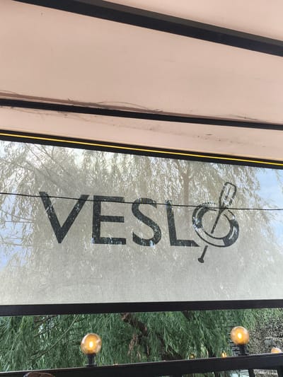 The image shows a sign with the text "VESLO" and a treble clef symbol, superimposed on a background of lush green trees. The sign appears to be made of black lettering on a white fabric material, possibly a canopy or a screen. The treble clef is stylized and has a playful, perhaps slightly abstract, design.

Below the sign, a railing is visible with several round, amber-colored lights attached to it, suggesting an outdoor setting, possibly a restaurant or cafe. The trees in the background are dense and leafy, with some branches visible. The overall impression is one of an outdoor space, likely a place for relaxation or entertainment, with a musical theme suggested by the treble clef. The lighting in the scene, with the warm glow of the lights and the diffused sunlight filtering through the trees, hints at either early morning or late afternoon. The image is taken from a slightly upward angle, looking towards the sign. No people are visible in the frame, and the focus is on the signage and the surrounding natural elements. The context provided indicates the location as Komsomolskoe, Kyrgyzstan.