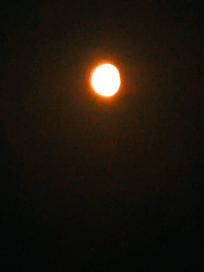 A full moon is visible in a dark, possibly smoggy sky. The moon is bright white with an orange halo and is the main subject of the image. The background is a dark, mottled brown and black. The image appears to be taken from an outdoor location in Shirokii Log, Russia, though no specific landmarks or people are visible.