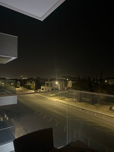 The image depicts an elevated nighttime view over a residential area in Latsia, Cyprus, from a balcony. The immediate foreground shows a white architectural structure on the left and a glass balcony railing extending across the lower half of the frame, with reflections visible on its surface. A section of a dark brown wicker chair and a light-colored surface are partially visible in the bottom right corner.
Below, a two-lane asphalt road with white dashed lane markings and diagonal white chevron patterns on the left runs horizontally, illuminated by several streetlights casting a yellowish glow. A prominent streetlight pole stands on the right side of the road. Multiple white or light-colored residential buildings, characteristic of modern architecture, are scattered throughout the midground, interspersed with dark vegetation. Several parked vehicles, including two dark-colored cars and one white car, are visible on the unpaved shoulder bordering the right side of the road.
In the background, a distant expanse of city lights extends towards the horizon, indicating a broader urban area. The sky is dark, with a faint crescent moon visible in the upper right quadrant. No people or active interactions are observable within the scene.