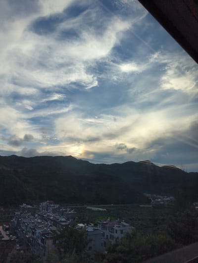 The image captures a panoramic view of a rural landscape at what appears to be sunset or sunrise, with dramatic cloud formations dominating the sky. The foreground is partially obscured by a dark, blurry element, possibly a window frame or railing. Below the sky, rolling, dark green hills form the middle ground, with a small, densely packed village nestled in the valley between them. The village consists of numerous multi-story buildings with grey roofs and white or light-colored walls. Some of the buildings have visible solar water heaters on their roofs. A road is visible winding through the valley, with a few cars parked and some movement along the road. The background is a silhouette of more hills against the bright sky. A faint rainbow or atmospheric optical phenomenon is visible in the sky above the hills. The overall impression is one of a quiet, mountainous region with a developing community. The location is identified as Mengzi City, China, a region known for its mountainous terrain and agricultural communities.