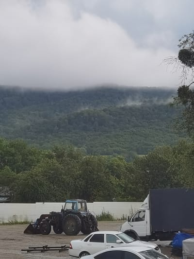 The image depicts an outdoor scene with a backdrop of mist-shrouded, heavily forested hills. In the foreground, on a dirt or gravel surface, are several vehicles. To the left, a blue tractor with a front loader is parked. To the right of the tractor is a white box truck with a dark blue tarp covering its cargo area. In front of these vehicles, two white sedans are visible. A white corrugated metal fence runs across the middle ground, with greenery behind it. The sky is overcast with clouds. The scene appears to be a yard or storage area for vehicles.