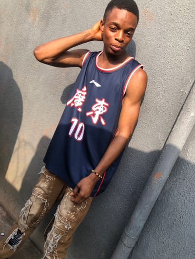 A young man poses in a sleeveless, dark blue basketball jersey with red and white trim and Chinese characters and the number 10 in white. He leans against a gray wall with a pipe, resting his head on his hand. He wears ripped, tan jeans with white paint splatters and a beaded bracelet. The photo is taken in bright sunlight, creating strong shadows.