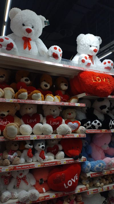 The image shows a retail display of stuffed teddy bears and heart-shaped pillows for sale, likely in a toy store or department store. The shelves are organized with various sizes and colors of plush toys, including large white teddy bears with red bows and paws, brown teddy bears, and a large red heart-shaped pillow with the word "LOVE" on it. Some of the toys have red hearts, and some have text that appears to be in Greek. The lighting suggests an indoor environment, possibly a supermarket or a dedicated toy shop. The display is stacked high with merchandise. The location is stated to be in Pylaia Municipal Unit, Greece. There are no people visible in the image.