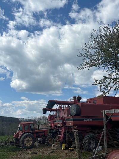 The image shows a Case IH combine harvester and a tractor parked near a chain-link fence. The machinery appears to be in a rural setting, possibly Presian, Bulgaria. The sky is visible with blue patches and white clouds. A tree with budding leaves is present in the upper left corner. The equipment is likely old or disused.