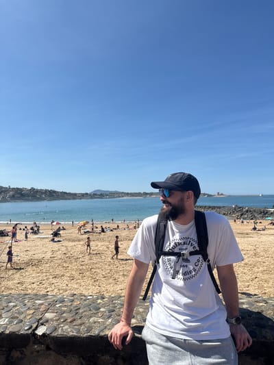 A man stands by a stone wall at a beach in Saint-Jean-de-Luz, France. He wears a white T-shirt, gray sweatpants, a black cap, and sunglasses, and a black backpack. The beach is crowded with people and the ocean is visible in the background. The sky is blue.