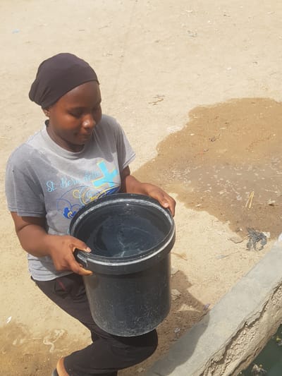 The photograph depicts a woman holding a large, black, cylindrical bucket filled with water. She is wearing a gray t-shirt with blue and gold designs, dark-colored leggings, and a dark head covering. The woman's posture indicates she is holding the bucket firmly. The environment appears to be an outdoor space with a combination of concrete, sand, and dirt surfaces. There's evidence of debris, including what looks like trash, scattered around. The location is indicated as Dutsinma A, Nigeria.