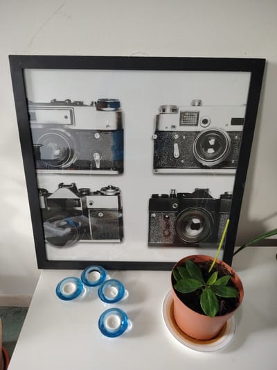 This image shows a framed picture of four vintage cameras on a white table. To the right of the framed picture, there is a small potted plant with green leaves. In front of the framed picture, there are four blue glass candle holders, each containing a white candle. The scene appears to be indoors, likely in a home or office setting. The location is not specified within the image itself or the provided crops.