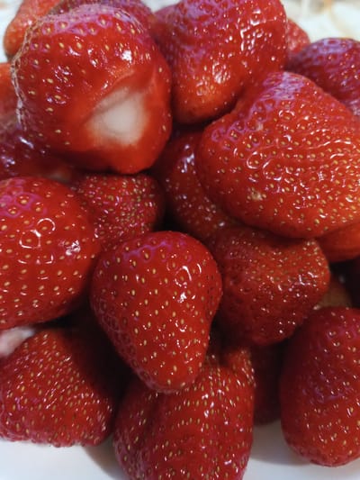 A close-up, overhead shot shows a pile of ripe, red strawberries on a white surface. The strawberries are whole and glistening, with visible seeds. One strawberry in the upper left corner is partially cut, revealing its white interior. The lighting highlights the glossy texture of the fruit. The location is not specified in the image, and no information is available about Suvorov, Russia.