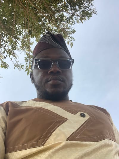 An adult male is depicted in an outdoor setting in Gindiri, Nigeria, viewed from a low-angle perspective. The subject, with a dark complexion and facial hair, wears clear-framed sunglasses with dark lenses, and a brown and beige striped traditional cap. He is dressed in a traditional garment featuring a light beige checkered pattern combined with solid brown fabric, forming geometric designs outlined by white stitching. A single dark, circular button is visible on the brown portion of the garment, near the right chest. Green foliage from a tree is partially visible in the upper left corner, set against a predominantly clear, light-colored sky. The subject looks directly towards the viewer.