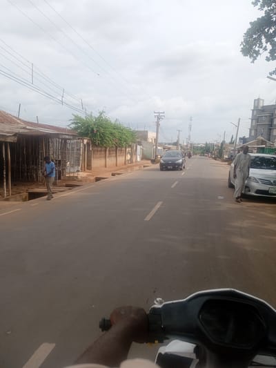 A low-angle, slightly blurry perspective from a motorcycle shows a street scene in Tundun Wada South, Nigeria. On the left, a single-story building with a corrugated iron roof has a barred entrance and displays a young man in a blue t-shirt and grey pants. Across the street, a black car is visible, and further down, a silver sedan is parked on the right, with a man in a light-colored robe standing next to it. The road is paved with white lines, and several power poles line the side of the road. The sky is overcast.