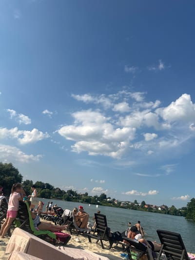 This media file captures a sunny day at a beach resort on a lake, likely in Dal'nie Prudishchi, Russia. The image is oriented vertically, with the sky dominating the left half and the beach and lake filling the right. Fluffy white clouds dot the clear blue sky, suggesting pleasant weather, likely midday due to the bright sunlight casting shadows and illuminating the scene.
The foreground features a sandy beach with several lounge chairs occupied by people. In the middle ground, the calm lake is visible, with a line of trees and buildings along the opposite shore, indicating a populated area or small town. Several individuals are seen relaxing in the chairs, some in swimwear, enjoying the warm weather. A few people are also visible in the water, perhaps swimming or wading. The overall mood seems relaxed and leisurely, typical of a summer day at a recreational spot.
One lounge chair in the foreground has a pink blanket with the word "ck" repeated on it. The scene depicts a typical summer day with people enjoying outdoor leisure activities by a body of water.