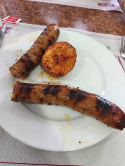 In the city of Polígono Industrial De Quart De Poblet, Spain, on a white plate, two grilled sausages and a slice of grilled potato are presented. The sausages are elongated and have prominent grill marks, with some areas appearing slightly charred. The potato slice is round with a golden-brown exterior and appears seasoned with a reddish spice, possibly paprika. The plate is slightly soiled with drippings from the food. The background suggests a restaurant or dining setting, with a textured countertop and what appears to be a placemat. The lighting is overhead and casts subtle shadows on the plate.