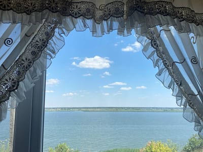 This image captures a scenic view of a body of water, likely a lake or large river, as seen through a window adorned with decorative curtains. The curtains are white and sheer, with elaborate lace trim featuring floral patterns and ruffled edges. They frame the view, creating a picturesque border.
The scene outside the window is dominated by the tranquil water, which occupies the lower half of the frame. Beyond the water, a distant shoreline is visible, lined with a stretch of greenery and what appear to be scattered buildings, suggesting a small settlement. The sky above is a vibrant blue, dotted with fluffy white clouds, indicating fair weather. The sunlight suggests it is daytime, likely late morning or early afternoon.
The overall impression is one of peacefulness and natural beauty. The presence of the curtains hints at an indoor setting, possibly a home or a room with a view, such as a guesthouse or a restaurant. The location context of Kyr'yakivka, Ukraine, adds a specific geographical reference to this serene landscape. There is no visible text in the image.