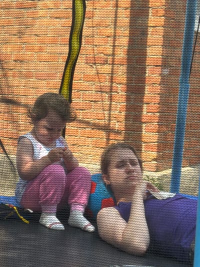 In Komsomolskiy, Russia, two individuals are captured in an outdoor setting, possibly a backyard, during daylight. A young child, seated, is wearing a sleeveless white top with a floral pattern and pink pants with white socks. The child is focused on something in their hands. Beside the child, an adult reclines, with their head resting on their hand. The adult is wearing a purple top. They are positioned on a dark-colored surface, likely a trampoline mat, which is part of a larger trampoline structure with a blue frame. The background consists of a textured, orange brick wall and a green and black striped element, possibly part of the trampoline's safety netting. Sunlight casts shadows across the scene, creating a dappled effect.