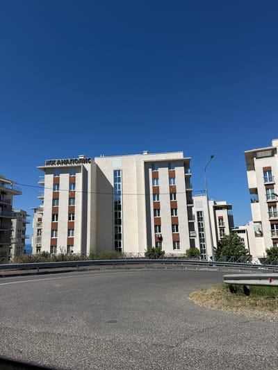 The image shows a street scene in Varvarovka, Russia, with several modern apartment buildings under a clear blue sky. In the foreground, a paved road curves to the right, with a metal guardrail separating it from a grassy area. The apartment buildings are white with dark red accent panels around the windows and balconies. One building has a sign with Cyrillic lettering that reads "ЭКАНАПОЛИС" on its upper facade. A streetlamp is visible between two of the buildings. The buildings appear to be residential, with multiple stories and balconies. The overall impression is one of a contemporary urban or suburban environment.