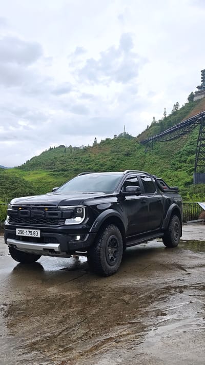 The image shows a black Ford Ranger Raptor pickup truck parked on a wet concrete surface. The truck has a license plate that reads "29K-179.83". The background features a green hillside with a metal structure resembling a cable car system. In the distance, there is a building or structure on the hilltop. The sky is overcast, suggesting cloudy weather. The scene is likely located in a mountainous or hilly area. Based on the visible elements, the location is consistent with the scenery found in Cat Cat Village, Vietnam, which is known for its mountainous terrain and cable car systems.