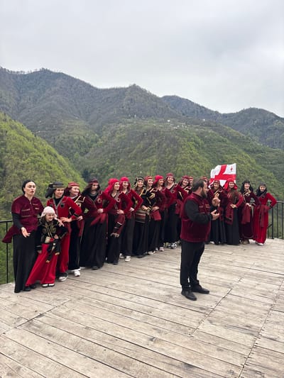 A wide-angle outdoor photograph captures a group of people posing on a wooden deck against a backdrop of mountains. The group consists of women and one man, all dressed in traditional Georgian clothing featuring red and black colors. The man appears to be leading or directing the group, gesturing with his hands. A Georgian flag is held aloft towards the back of the group. The deck is made of light-colored wooden planks and stretches across the lower portion of the frame. Behind the group, a mountain range covered in green vegetation rises towards a cloudy sky. The city of Kveda Chkhutuneti, Georgia is not visible in this image.