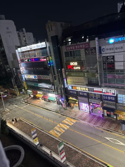 This is a night-time, high-angle, slightly tilted, vertical shot of a bustling street in Cheongju-si, South Korea. The street is lined with multi-story buildings adorned with numerous illuminated signs advertising various businesses. Neon lights in vibrant colors like blue, pink, red, and yellow illuminate the storefronts and the street itself.
The buildings house a variety of establishments including a PC cafe ("BOB PREMIUM PC CAFE"), a dental clinic ("김기훈치과", "미소치과"), a rehabilitation clinic ("88재활의학과의원"), a gym ("몸조아GYM"), a tutoring academy ("강남투탑 영수학원"), and a beauty salon ("엘린헤어"). Some signs indicate late-night or emergency services, such as "야간진료" (nighttime treatment). The road in front of the buildings has yellow lines and a pedestrian crossing with yellow and white stripes. There are a few individuals visible on the sidewalk. The overall impression is one of a lively urban commercial district at night.