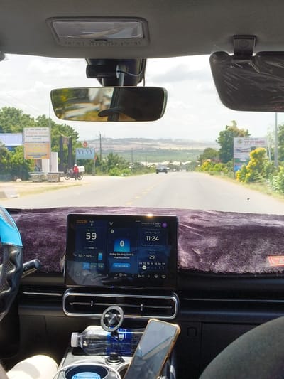 The image is a first-person perspective from inside a car traveling on a road in Hòa Thắng, Vietnam. The car's dashboard is visible, featuring a large touchscreen display showing the car's speed at 59 km/h, GPS information, and weather data indicating an outside temperature of 29.5°C. A steering wheel with a blue cover is on the left. Outside the car, a two-lane asphalt road stretches ahead, lined with trees and vegetation. Signs for "THANH TUNG" and "VY VY FARM" are visible on the roadside. In the distance, hills and a partly cloudy sky are present. A motorcycle with a rider wearing a white helmet and carrying a red box is moving away from the camera on the side of the road. The interior of the car also contains a water bottle and a smartphone resting on a holder.