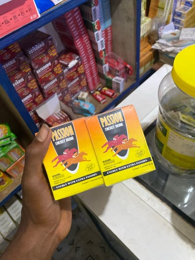 This is a close-up, eye-level shot of a person's hand holding two yellow and orange boxes of "Passion Energy Drink" in front of a shelf stocked with various medicines and packaged goods. The boxes are stacked vertically, with the top one slightly obscuring the bottom one. The "Passion Energy Drink" boxes feature an illustration of three horses in red and orange hues and prominent text in a dark font. Visible text on the boxes includes "PASSION ENERGY DRINK," "SUGAR FREE," "ENERGY WITH EXTRA PASSION," "NO ALCOHOL," "NO PRESERVATIVES," and regulatory information like "NAFDAC Reg. No. 01-8933" and "FDB/Dk 09-128". The text "Content: 6 sachets @4g" is also visible.

Behind the energy drink boxes, a blue shelf is filled with rows of red and white packaged items, many of which have "OSTEOKRIS" printed on them. Further in the background, out of focus, are more products, a glass jar with a yellow lid, and what appears to be a counter or another shelf. The lighting suggests an indoor setting, likely a store or pharmacy, possibly during daylight hours given the ambient light. The hand holding the boxes is dark-skinned, with a thumb and forefinger visible, appearing to be positioned to show the product. The overall impression is of a retail environment where these energy drinks are being presented or purchased. The location context provided is Jere South, Nigeria.